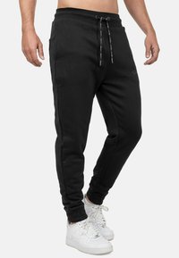 Zwarte sweatpants gemaakt van zachte stof, met een elastische tailleband met trekkoord, taps toelopende pijpen en zijzakken. Gecombineerd met witte sneakers.