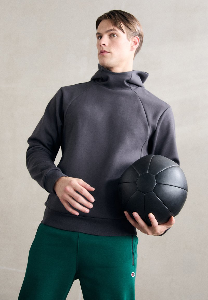 Felpa grigia con cappuccio, colletto alto e maniche raglan, abbinata a pantaloni tuta verdi. Il modello tiene un pallone da fitness nero.
