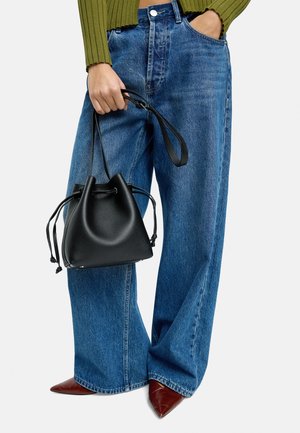 Czarna skórzana torba typu bucket z zapięciem na sznurek, trzymana w jednej ręce. Jasnoniebieskie dżinsy o szerokich nogawkach z wysokim stanem oraz czerwone czółenka ze spiczastym noskiem.