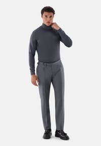 Grauer Rollkragenpullover, maßgeschneiderte graue Hose mit subtiler Textur und schwarze Slipper. Das Modell posiert mit einer Hand am Kragen.