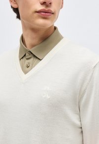 Άνδρας με ανοιχτό μπεζ πουλόβερ V λαιμόκοψης πάνω από πουκάμισο ελιάς με κουμπιά, εστιασμένος στο άνω μέρος του στήθους και το κάτω μέρος του προσώπου σε απλό φόντο.