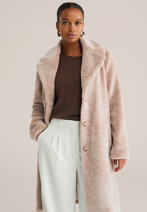 Cappotto in pelliccia sintetica rosa blush con colletto a scialle, bottoni frontali e tessuto testurizzato; indossato sopra una maglia marrone e pantaloni a righe verde chiaro.
