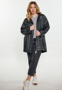 Chaqueta de lluvia negra y brillante con capucha, botones a presión, dos bolsillos laterales; llevada sobre una camiseta de rayas y combinada con jeans oscuros y zapatillas.