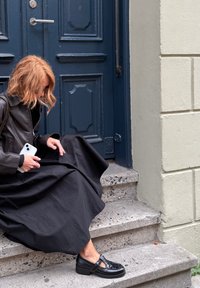 Black long skirt drapes over gray stone steps, paired with shiny black platform shoes featuring a buckle. A dark leather jacket completes the look.
