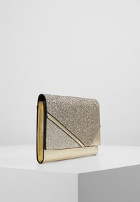 Zlatá a stříbrná třpytivá kabelka ve tvaru obálky s metalickými akcenty, stojící vzpřímeně na bílé ploše na pozadí jednoduché světle šedé barvy.