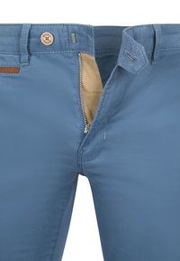 Helleblaue Hosen mit einem Reißverschluss, beige Innenfutter und braunen Lederakzenten am Bund. Glatte Stoffstruktur.