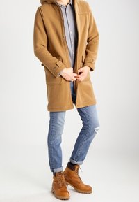 Osoba zapinająca guziki na camelowym płaszczu, ubrana w niebieskie podwinięte dżinsy i beżowe sznurowane buty, na jednolitym tle.