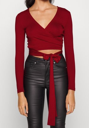 Kobieta ubrana w długim rękawem głęboko czerwoną bluzkę crop top z wiązaną kokardą w talii, połączoną z czarnymi, skórzanymi spodniami z wysokim stanem.