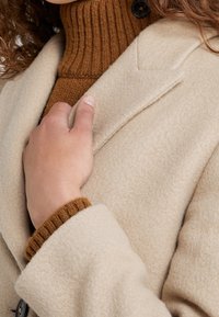 Beiger Wollmantel mit strukturiertem Finish, mit schmalem Revers und Knopfdetails. Brauner Ripp-Turtleneck sichtbar darunter.