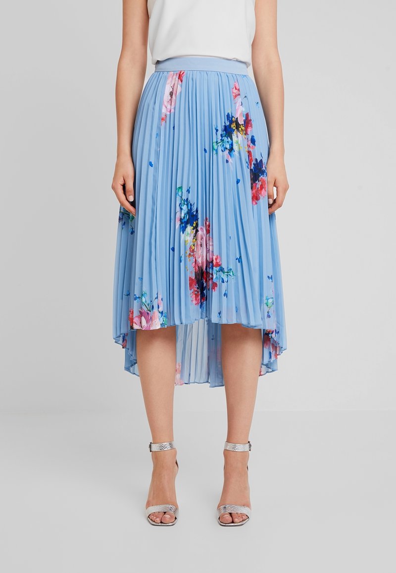 Femme portant une jupe midi plissée bleu clair à imprimés floraux et des talons argentés à bride à la cheville, debout devant un fond gris uni.