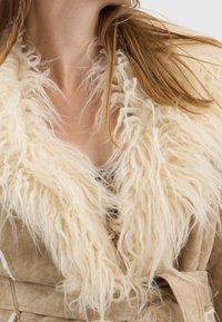 Cappotto in suede beige con un colletto in pelliccia sintetica bianca, spesso e morbido. Il colletto presenta fibre lunghe e texture. Il cappotto è dotato di una vita regolabile e linee diagonali.