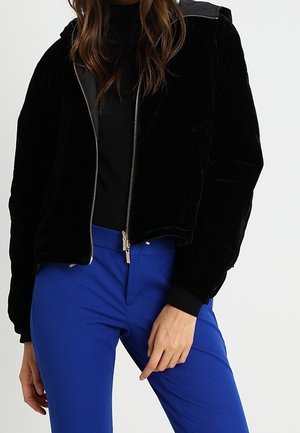 Frau trägt eine schwarze Jacke mit Reißverschluss und Kapuze sowie leuchtend blaue Hose, steht mit einer Hand nahe der Hüfte vor einem schlichten Hintergrund.