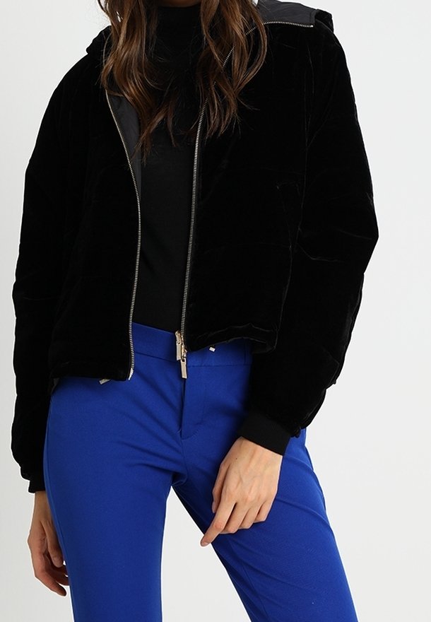 Femme portant une veste noire zippée avec une capuche et un pantalon bleu vif, debout avec une main près de sa hanche sur un fond uni.