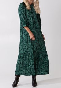 Vestido largo verde en capas con estampado de paisley, con escote redondo y mangas tres cuartos. Combinado con botas negras hasta el tobillo.