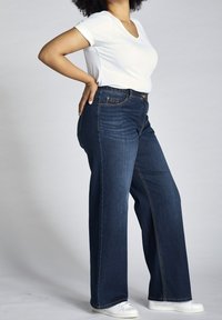 Mörkblå vida jeans med sydda fickor, kombinerade med en åtsittande vit T-shirt med V-ringning och vita sneakers. Slät denimtextur synlig.