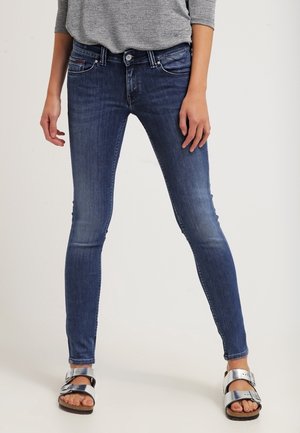 Personne portant un jean skinny bleu foncé, une chemise grise à manches longues et des sandales argentées à boucle, debout les jambes légèrement écartées.