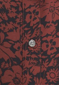 Červená košile s černým květinovým vzorem, s kovovou knoflíčkem a kontrastním stehem podél švu. Hladká textilie.