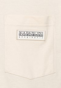 Camiseta de algodón color crema con un bolsillo cuadrado de parche y una etiqueta cosida que muestra "NAPAPIJRI geographic" en negro.