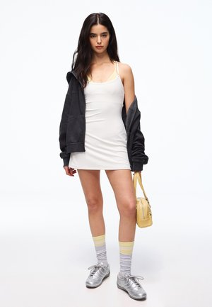 Młoda kobieta w białej sukience na ramiączkach, czarna kurtka zsunięta z ramion, szare i żółte skarpety, srebrne buty, trzyma żółtą torebkę, stoi na tle białego tła.