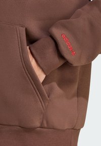 Sweatshirt marrone con tasca; polsini a coste; logo "adidas" ricamato in rosso sulla manica sinistra; tessuto morbido e strutturato.