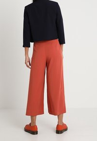Veste courte bleu marine, pantalon large couleur rouille et chaussures orange. Tissu léger et lisse avec une coupe ajustée et des détails subtils sur la veste.