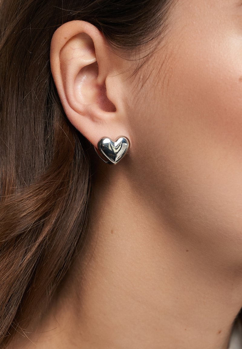 Sølv hjerteformede øreringe med en poleret overflade, båret i øret, som fremviser glatte konturer og reflekterende overfladedetaljer.