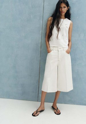 Femme aux longs cheveux foncés porte un haut blanc sans manches et un pantalon blanc ample et court, debout les mains dans les poches contre un mur bleu.