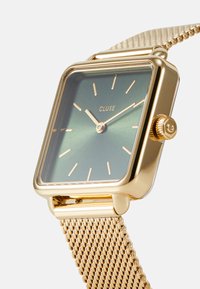 Montre en or carrée avec un cadran dégradé vert, marqueurs d'heure minimalistes et un bracelet en maille. Dispose d'une couronne sur le côté pour les réglages.