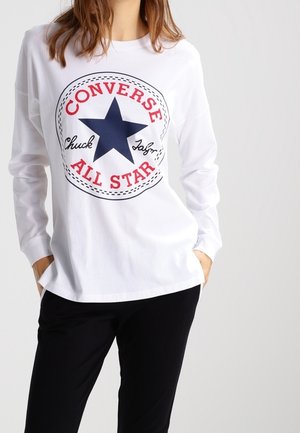 Persoon die een wit overhemd met lange mouwen draagt met een groot Converse All Star-logo en een zwarte broek, handen in de zakken.