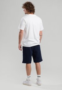 Valkoinen puuvillapaita lyhythihaisilla hihoilla, yhdistettynä tummansinisiin shortseihin ja valkoisiin lenkkitossuihin. Näkyvät raidat nilkkasukissa.