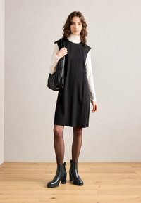 Vestido negro sin mangas con corte relajado, combinado con un suéter de cuello alto blanco. El modelo lleva botas de tacón negras hasta el tobillo y un bolso negro texturizado.