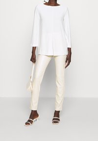 Biała bluzka z długim rękawem i okrągłym dekoltem, w połączeniu z błyszczącymi, kremowymi spodniami slim-fit. Modelka trzyma także małą, jasną torebkę.
