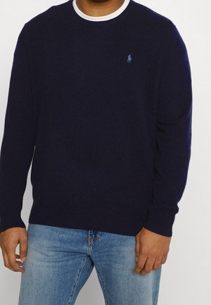 Man draagt een marineblauwe trui met ronde hals en een klein logo op de borst en lichtblauwe spijkerbroek, staand tegen een effen achtergrond.