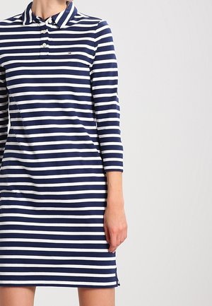 Robe rayée bleu marine et blanche avec un col à revers, des manches longues et des boutons sur la patte de boutonnage. Présente des fentes latérales pour faciliter les mouvements.