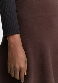 Geweven zwart langemouwen top met ribbeltextuur, gecombineerd met een strakke bruine stoffen rok. Close-up van de hand die op de rok rust.