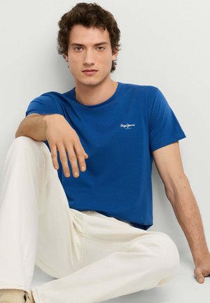 Joven de pie vistiendo una camiseta azul de manga corta, pantalones color beige claro y zapatos beige sin cordones, sobre un fondo liso y claro.