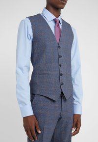 Man wearing a blue checked waistcoat and matching trousers with a light blue shirt and purple textured tie.