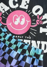 Svart t-shirt med en stor rosa smiley-grafik och vit text. Kombineras med randiga shorts i flerfärgade nyanser av blått och lila.