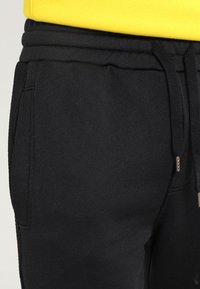 Černé tepláky z texturované látky s šňůrkou v pase a postranními kapsami. Pas je pružný žebrovaný pro pevné uchycení.