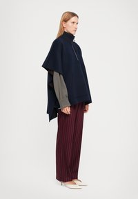 Poncho navy con colletto alto, chiusura con zip e tasca frontale, indossato sopra una maglietta grigia, abbinato a pantaloni borgogna a righe e scarpe beige.