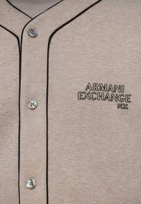 Camicia button-up in cotone grigia con piping neri e logo Armani Exchange ricamato in nero. Presenta un bottone argentato al colletto.