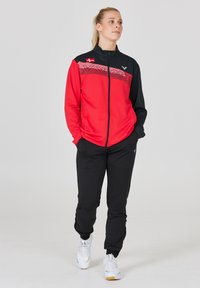 Rød og sort atletisk jakke med lynlås foran og dansk flag detalje; parret med sorte bukser og hvide sneakers. Glat stof og tætsiddende design.