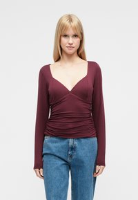 Burgundy long-sleeve top with a deep V-neck, ruched detailing at the waist, and a fitted design, paired with light blue denim jeans.