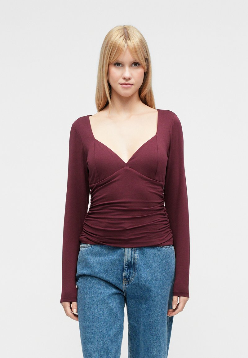 Burgundy long-sleeve top with a deep V-neck, ruched detailing at the waist, and a fitted design, paired with light blue denim jeans.