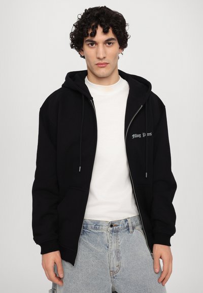 Hanorac negru cu fermoar, cu un buzunar frontal și șnururi, purtat peste un tricou alb. Prezintă un text brodat subtil pe piept.