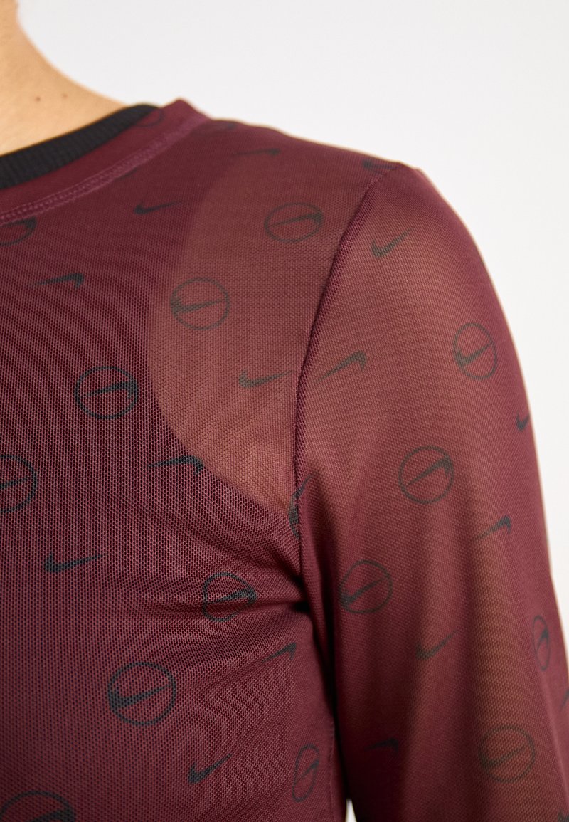 Bordeaux sporttop met meshstof en een herhaald Nike-logo patroon. De halslijn is zwart en het heeft aansluitende lange mouwen.