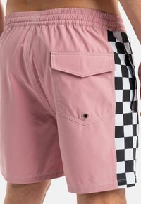 Bañador rosa con cintura elástica, bolsillo lateral y un patrón de tablero de ajedrez en blanco y negro en el lado izquierdo. Textura suave.