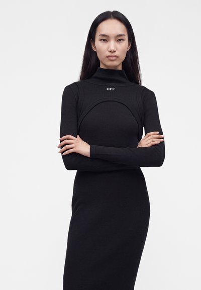 Femme aux longs cheveux noirs portant une robe noire ajustée avec un col montant et les bras croisés sur un fond clair uni.