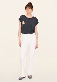 Graues Kurzarm-T-Shirt mit floraler Stickerei, kombiniert mit weißen Schlagjeans und grauen Ballerinas vor einem neutralen Hintergrund.