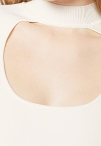 Gros plan d'une personne portant un pull blanc avec un large col et un détail découpé sous le col.